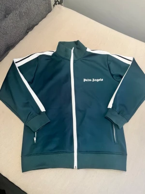 Grön track jacket från Palm Angels - Snygg grön track jacket från Palm Angels med vita ränder längs ärmarna och vit dragkedja framtill. Jackan har hög krage, långa ärmar och logga broderad på bröstet. Perfekt för en sportig och trendig look.