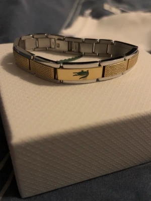 Lacoste armband - Snyggt armband från Lacoste i guldfärgad metall. Stilren och modern design som passar både till vardag och fest. Armbandet är i mycket fint skick och kommer komplett med originalförpackning, ask, tygpåse och etikett – perfekt även som present. Finns extra länkar om den inte passar pris går diskutera ny pris 1200kr