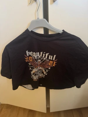 Svart croppad t-shirt med tryck - Cool svart croppad t-shirt med tryck framtill som har en örn, texten 'beautiful', '2021' och rockiga detaljer. Perfekt för dig som gillar edgy streetstyle och vill sticka ut. T-shirten har korta ärmar och en avslappnad passform.