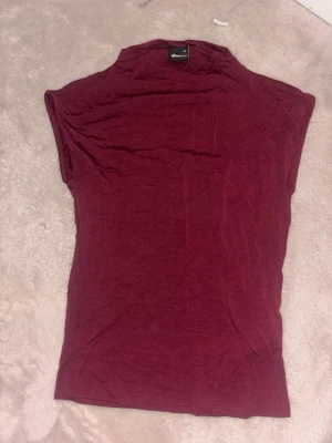 Vinröd polotopp från Gina Tricot - Vinröd polotopp från Gina Tricot i mjukt material. Toppen har en hög krage och är ärmlös med en slim passform. Perfekt att styla med jeans eller kjol för en trendig look.