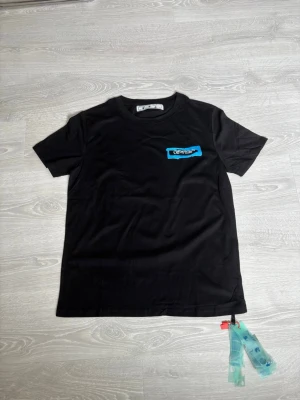 Svart Off-White t-shirt med blå detalj - Snygg svart t-shirt från Off-White med korta ärmar och rund hals. På bröstet finns en blå detalj med Off-White-loggan. T-shirten har även de klassiska Off-White-taggarna i nederkant. Perfekt för dig som gillar streetwear och vill sticka ut. Stor i storlek!!