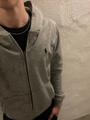 Grå zip hoodie från Polo Ralph Lauren - Grå hoodie från Polo Ralph Lauren med dragkedja framtill och klassisk huva. Ikoniska loggan broderad på bröstet. Mjuk bomullsblandning och två fickor fram. 