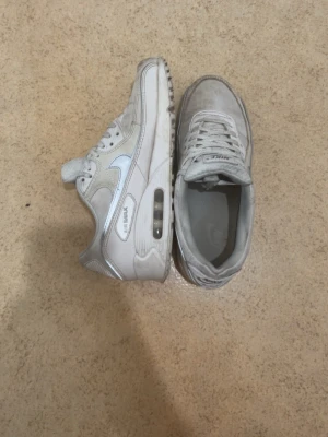 Vita Nike Air Max 90 sneakers - Säljer ett par klassiska vita Nike Air Max 90 sneakers med synlig Air-enhet i sulan. Skorna har en kombination av läder och mesh i ovandelen, samt den ikoniska swoosh-loggan på sidan. Perfekta för dig som gillar stilrena och tidlösa sneakers.