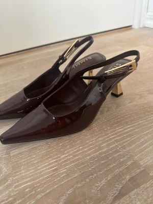 Bruna lackpumps med gulddetalj - Intressekoll på ett par mörkbruna pumps från Simmi London i glansigt lackmaterial. Skorna har spetsig tå, öppen häl med justerbar rem och snygg guldfärgad detalj vid spännet. Ordinarie pris 429 kr. Endast prövade. 