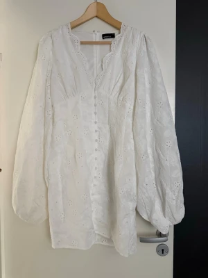 Vit kort klänning från Gina Tricot - Söt vit kort klänning från Gina Tricot med broderade blommor, knappar framtill och ballongärmar. Klänningen har v-ringad hals med spetsdetaljer och är perfekt för sommarens alla tillfällen. Den är helt oanvänd, pågrund av att den va lite för stor mig mig som vanligtvis har s/xs.