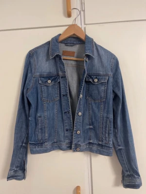 Klassisk blå jeansjacka - Snygg blå jeansjacka i klassisk modell med två bröstfickor med lock och knappar, samt sidofickor. Jackan har silverfärgade knappar framtill och vid ärmsluten. Perfekt att slänga över en hoodie eller t-shirt för en avslappnad look.