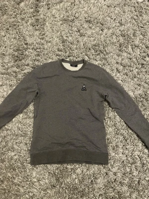 Grå sweatshirt från jlindeberg - Säljer en stilren grå sweatshirt från J Lindeberg med rund halsringning och ett diskret svartvitt märke på bröstet. Tröjan har långa ärmar och ribbade muddar vid ärmslut och nederkant. Perfekt för en avslappnad look.