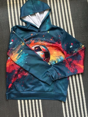 Färgstark hoodie med fotbollsmotiv - Säljer en cool hoodie med huva i mörkblått och färgstarka splash-effekter i rött, orange och turkost. Mitt på bröstet finns ett stort fotbollsmotiv. Hoodien har ribbade muddar och är tillverkad i ett mjukt, syntetiskt material. Perfekt för dig som gillar sportig stil. Storlek 160