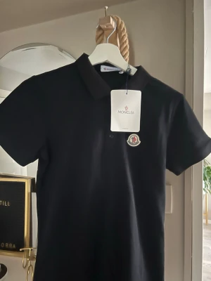 Svart Moncler pikétröja - Snygg svart pikétröja från Moncler med klassisk krage och korta ärmar. Tröjan har Monclers ikoniska logotyp på bröstet och är tillverkad i mjuk bomull. Perfekt för en clean och stilren look.