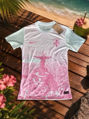 Nike t-shirt med Rio de Janeiro-motiv - Unik t-shirt från Nike med korta ärmar och ett stort rosa motiv av Kristusstatyn i Rio de Janeiro. T-shirten är vit med ljusblå toner och har Nike-logga på bröstet. Tillverkad i ett lätt och bekvämt material, perfekt för sommardagar.
