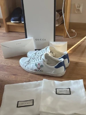 Gucci Tennis skor - Säljer ett par vita Gucci sneakers med blå detaljer på hälen och broderad tennisracket-design på sidan. Skorna har klassisk låg profil, vita snören och är tillverkade i läder. Medföljer originalförpackning och dustbags. Hör av er vid andra frågor 🙏
