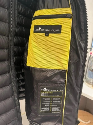 Svart pufferjacka Moose Knuckles - Snygg svart pufferjacka från Moose Knuckles med quiltad design och gul meshficka på insidan. Jackan har dragkedja framtill och står ut med sin lätta och smidiga känsla. Perfekt för kalla vinterdagar och har en modern, sportig look. Finns en reva.