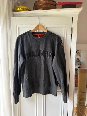 Mörkgrå sweatshirt från Parajumpers - Mörkgrå sweatshirt från Parajumpers med rund halsringning och stor logga broderad över bröstet. Tröjan har långa ärmar, ribbade muddar och en patch på ena ärmen. 