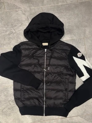 Svart hoodie från Moncler med dragkedja - Snygg svart hoodie från Moncler med vadderad framsida och stickade ärmar. Dragkedja framtill, två fickor med knapp och stor huva. På vänster ärm finns ett vitt mönster och Moncler-logga. Perfekt för dig som vill ha en exklusiv streetwear-look. Även NFC funkar. Priset går SJÄLVKLART att diskuteras vid bra affär! 