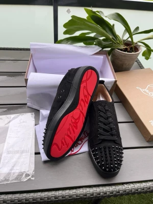 Svarta Louboutin sneakers med nitar - Säljer ett par svarta sneakers från Christian Louboutin i mocka med svarta nitar på tån och den ikoniska röda sulan. Skorna har svarta skosnören och en låg siluett som ger en clean och edgy look. Perfekta för dig som vill sticka ut med exklusiva detaljer.