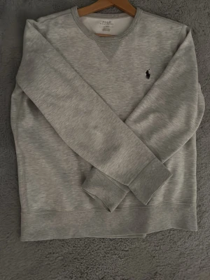 Grå sweatshirt från Polo Ralph Lauren - Snygg grå sweatshirt från Polo Ralph Lauren med klassisk rund halsringning och broderad logga på bröstet. Tröjan har långa ärmar, ribbade muddar och är gjord i mjukt bomullsmaterial. Perfekt för en avslappnad och stilren look. Passar folk från 170-180  ej använd inköpt på NK förra året 