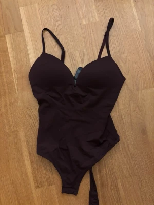 Vinröd body med tunna axelband - Säljer en mörkröd body med tunna, justerbara axelband och vadderade kupor. Bodyn har en djup urringning och är tillverkad i ett stretchigt material som formar sig efter kroppen. Perfekt för att skapa en snygg siluett under kläder.