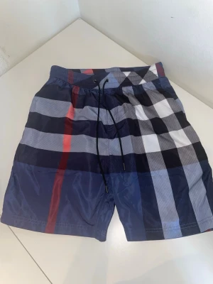 BURBERRY badbyxor  - Snygga badbyxor med klassiskt rutmönster i marinblått, vitt, svart och rött. De har resår i midjan med snörning och en bakficka. Perfekta för stranden eller poolen och ger en stilren look. Materialet känns lätt och snabbtorkande. Helt nya, har bara testat dem men har Tyvär slarvat bort lappen.