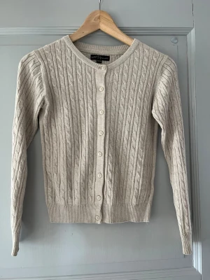 Beige kabelstickad kofta  - Kabelstickad beige kofta med rund halsringning och knäppning framtill. Långa ärmar och ribbade muddar vid ärmslut och nederkant. Perfekt för lager-på-lager och enkel att matcha med olika outfits.