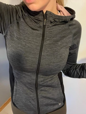 Mörkgrå träningsjacka med huva - Mörkgrå melerad träningsjacka från SOC/Stadium med hel dragkedja och huva. Jackan har svarta detaljer längs sidorna, rosa i luvan och är tillverkad i ett stretchigt funktionsmaterial som andas. 