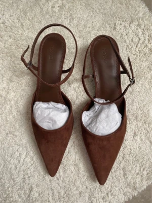 Bruna pumps i mocka från NA-KD - Snygga bruna pumps från NA-KD i mjuk mocka med spetsig tå och smal klack. Skorna har en elegant rem runt ankeln med justerbart spänne. Perfekta för dig som vill ha en stilren och trendig look.