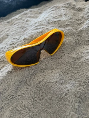 Gula futuristiska solglasögon - Coola solglasögon med bred, gul plastbåge och mörka linser. Glasögonen har en futuristisk, sportig form som sticker ut och ger en unik look. Perfekta för dig som vill ha något annorlunda och vågat till sommaren.