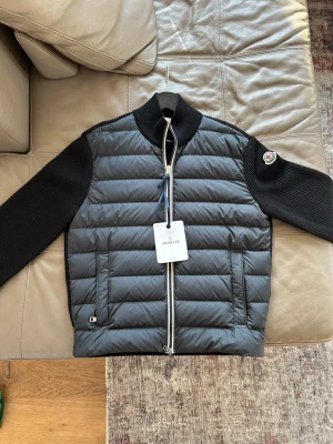 cardiganjacka från moncler köpt på NK Göteborg, kvitto finns  - Moncler cardiganjacka i svart med quiltad dunfront och ribbstickade ärmar och rygg. Jackan har en krage, dragkedja framtill, två fickor och Moncler-logga på vänster ärm. Materialmix av dun och stickat ger en modern och sportig look.