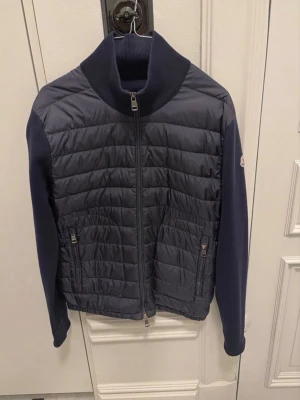 Mörkblå cardiganjacka från Moncler - Snygg mörkblå cardiganjacka från Moncler med quiltad front och stickade ärmar. Jackan har hög krage, dragkedja framtill och två fickor med dragkedja. Perfekt för lager på lager och en stilren look. ÄKTA 