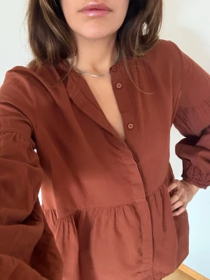 Bohemisk rostbrun blus – luftig modell med volymärmar | A Part of the Art - Elegant och luftig blus i en varm rostbrun ton från A Part of the Art. Den har en avslappnad bohemisk siluett med vackra volymärmar, lätt rynkning under bysten och en feminin knappdetalj framtill.  Tyget faller mjukt och ger en luftig, naturlig känsla – perfekt både till vardag och mer uppklädda tillfällen.