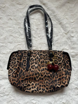 Leopardmönstrad handväska med körsbärnyckelring - Cool handväska med leopardmönster i brunt och svart. Väskan har svarta handtag och en glittrig körsbärsberlock i rött och guld. Perfekt statement piece för dig som gillar djurmönster och unika detaljer. Materialet ser ut att vara konstläder eller textil.