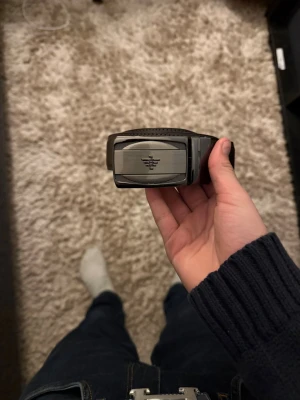 Svart bälte med metallspänne från Armani - Snyggt svart bälte från Armani med ett stilrent metallspänne i silverfärg och logga framtill. Bältet är gjort i skinn och passar perfekt till jeans eller kostymbyxor. Enkel och modern design som lyfter din outfit.