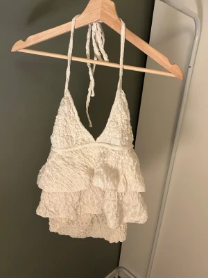 Vit volang topp med halterneck - Supersöt vit topp med halterneck och flera lager volanger. Smockad struktur i tyget ger en trendig look och toppen knyts i nacken. Perfekt för sommaren och passar till både jeans och kjol.
