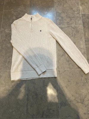 Morris tröja med halv dragkedja - Snygg beige stickad tröja med halv dragkedja och hög krage. Tröjan har ett diskret mörkblått broderat emblem på bröstet och ribbade muddar vid ärmslut och nederkant. Perfekt för lager på lager och enkel att matcha.