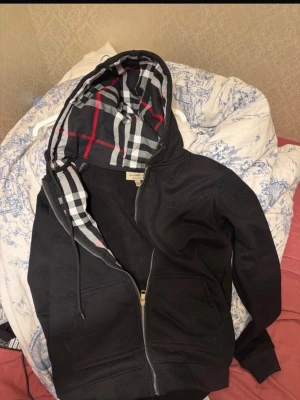 Svart hoodie med dragkedja Burberry - Säljer nu en svart hoodie med dragkedja från Burberry. Klassisk huva med Burberrys rutiga mönster i svart, vitt och rött på insidan. Två fickor framtill och justerbar dragsko i huvan. Perfekt för en stilren och avslappnad look. Skriv vid frågor, tveka inte! Pris kan även diskuteras:)