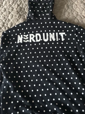 Svart prickig hoodie från Nerdunit - Säljer en svart hoodie från Nerdunit med vita prickar över hela och stor vit logga på ryggen. Hoodien har huva och en avslappnad passform, perfekt för dig som gillar streetwear och vill sticka ut lite extra. Materialet känns mjukt och bekvämt.
