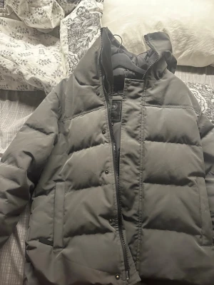 Mörkgrå pufferjacka från Canada Goose - Säljer en mörkgrå pufferjacka från Canada Goose med hög krage och dragkedja framtill. Jackan har två sidofickor och en patch med logga på ärmen. Perfekt för kalla dagar och riktigt varm tack vare det tjocka materialet.