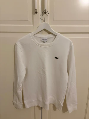 Vit sweatshirt från Lacoste - Vit sweatshirt från Lacoste med klassisk rund hals och det ikoniska krokodilmärket broderat på bröstet. Tröjan har långa ärmar och ribbade muddar vid ärmslut och nederkant. Perfekt för dig som gillar stilren och enkel design.
