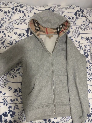 Grå hoodie med dragkedja Burberry - Snygg grå hoodie från Burberry med dragkedja framtill och klassiskt rutigt foder i huvan. Mjuk bomullsmix, två fickor fram och ribbade muddar. Perfekt för en chill och stilren look.