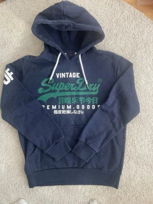 Mörkblå Superdry hoodie med tryck - Snygg mörkblå hoodie från Superdry med grönt och vitt tryck på bröstet och japanska tecken. Klassisk huva med vita snören och känguruficka framtill. Mjuk insida och ribbade muddar. Perfekt för en avslappnad streetwear-look.