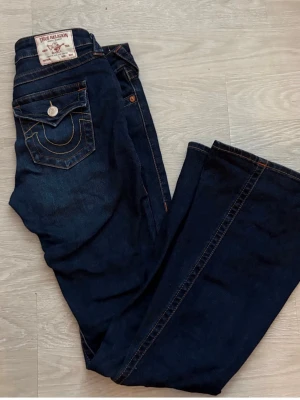 Mörkblå joey low Rise jeans True Religion 27 (28)/34  - Säljer ett par mörkblå bootcut jeans med log midja ”joey low rise” från True Religion med klassiska kontrastsömmar och ficklock bak. Storleken upplevs mer som 28/34 än 27/34.  Jeansen har låg midja och silverfärgade detaljer. Perfekta för dig som gillar en avslappnad men ändå trendig stil 