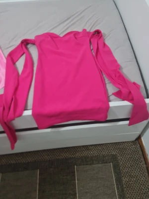 Rosa kort klänning från JI - Supersnygg rosa kort klänning från JI med axelbandslös design och långa knytband vid axlarna. Klänningen har en figurnära passform och är tillverkad i ett mjukt syntetmaterial som känns bekvämt mot huden. Perfekt för dig som vill sticka ut med färg.