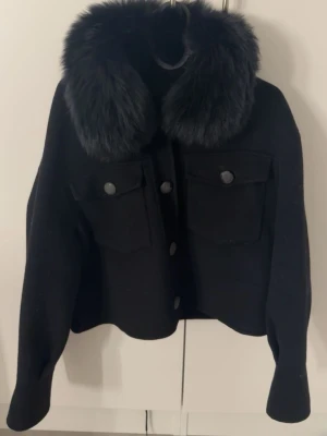 Svart pälsjacka med stor krage - Snygg svart pälsjacka med stor fluffig fuskpäls-krage. Jackan har två stora bröstfickor med lock och knappar framtill. Kortare modell med raka ärmar och stilrena detaljer. Perfekt för dig som vill ha en trendig och varm jacka i vinter. Knapparna är lite slitna men inget man tänker på. Köpt för ungefär 2300