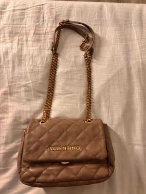 ”Smutsrosa” axelväska från Valentino - Snygg ”smutsrosa” axelväska från Valentino med quiltat mönster och guldfärgad kedja. Väskan har ett elegant guldigt logotyp på framsidan och en praktisk ficka på baksidan. Materialet är mjukt och lyxigt, perfekt för dig som vill ha en trendig och stilren look.