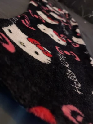 Hello Kitty pyjamasbyxor svart/rosa - Mysiga pyjamasbyxor i svart fleece med Hello Kitty-motiv och rosa detaljer. Byxorna har ett heltäckande tryck med Hello Kittys ansikte och rosetter, samt texten 'Hello Kitty' i vitt. Perfekta för chillkvällar hemma.