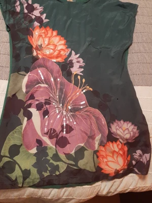 Blommig topp med paljettdetaljer från Next - Unik topp med stort blommönster i lila, orange och grönt på mörk botten. Framsidan har paljettdetaljer som ger extra glans. Toppen är ärmlös och har en mjuk, följsam passform. Perfekt för dig som vill sticka ut med färg och mönster. Jag tror är det större än storlek UK 10/EU 38. Mått på bilderna.