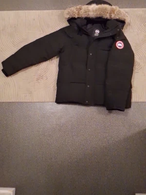 Svart dunjacka från Canada Goose - Säljer en svart dunjacka från Canada Goose med huva och avtagbar pälskant. Jackan har klassisk logga på bröstet, dragkedja och knappar framtill samt flera fickor. Perfekt för kalla vinterdagar och riktigt varm.