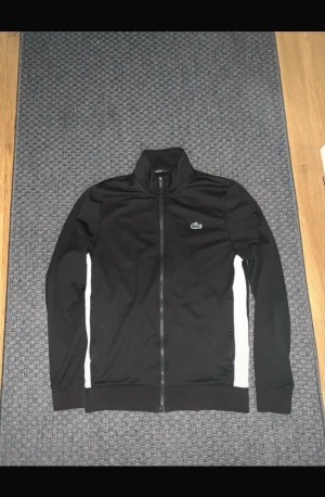 Svart Kofta från Lacoste Sport - Svart Kofta från Lacoste Sport med vita paneler längs sidorna och klassisk krokodillogga på bröstet. Tröjan har hög krage, hel dragkedja och ribbade muddar. Perfekt för en sportig och stilren look. 