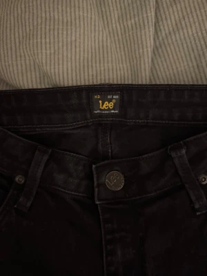 Svarta raka jeans från Lee - Säljer ett par svarta jeans från Lee med raka ben. Jeansen har knappgylf med Lee-logga och är tillverkade i ett mjukt denimtyg som sitter snyggt.