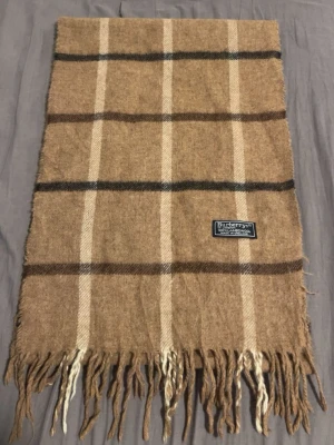 Beige rutig halsduk från Burberrys - Klassisk beige halsduk från Burberrys med rutigt mönster i brunt, svart och vitt. Tillverkad i 100% lammull och har fransar i båda ändar. Märkt 'Made in England' och har en ikonisk Burberrys-label. Perfekt accessoar för kyliga dagar.