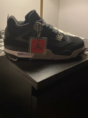 Svarta Air Jordan 4 Retro sneakers - Säljer ett par svarta Air Jordan 4 Retro sneakers med vita och grå detaljer. Skorna har meshpaneler, plastdetaljer vid snörningen och en synlig Air-enhet i sulan. Klassisk Jumpman-tag hänger vid sidan. Perfekta för dig som gillar streetwear och ikoniska modeller.